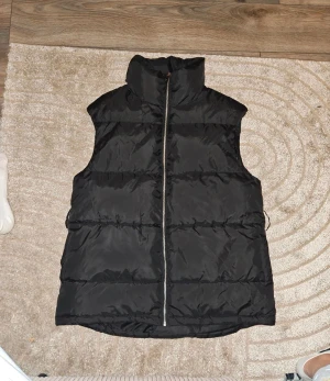 Svart dunväst från NLY Trend - Svart dunväst från NLY Trend i storlek 38. Västen har hög krage, dragkedja framtill och praktiska fickor på sidorna. Den är quiltad och har en loose passform, perfekt för lager-på-lager under kyliga dagar. Materialet är syntet och polyester.