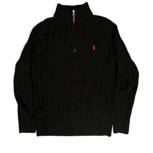 Svart kabelstickad tröja Polo Ralph Lauren - Svart kabelstickad tröja från Polo Ralph Lauren med halv dragkedja och röd broderad logga på bröstet. Tröjan har ribbad krage, muddar och är långärmad. Perfekt för dig som gillar klassisk stil med en modern touch.