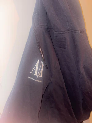Svart hoodie från Armani Exchange - Svart hoodie från Armani Exchange med stor vit logga på ärmen och snygga detaljer på snörningen. Tröjan har huva och är tillverkad i mjuk bomull, perfekt för chill dagar. Klassisk passform och stilren look som funkar året runt.