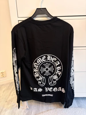 Svart långärmad Chrome Hearts tröja - Svart långärmad tröja från Chrome Hearts med vita tryck på ryggen, bröstet och båda ärmarna. Snygga detaljer med logotyp och Las Vegas-tryck, samt CH-bokstäver vid nederkanten. Tröjan har rund hals och är tillverkad i mjuk bomull. Pris diskuteras vid snabb affär.