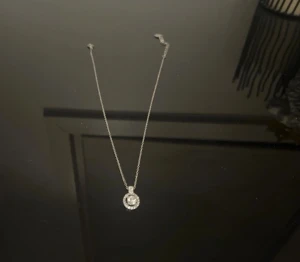 Silverfärgat halsband med glittrig cirkel - Ett stilrent halsband i silverfärg med en tunn kedja och ett runt hänge dekorerat med gnistrande stenar. Hänget har en cirkelformad design som ger en elegant och modern känsla. Perfekt för dig som gillar diskreta men ändå iögonfallande smycken.från montini 