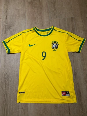 Brasilien Ronaldo #9 fotbollströja Nike - Gul Brasilien fotbollströja från Nike med gröna detaljer, nummer 9 och Ronaldo på ryggen. Tröjan har korta ärmar, rund hals och officiellt CBF-emblem på bröstet. Tillverkad i lätt polyester med NikeFit-teknologi för skön känsla på planen.   Storlek S/M