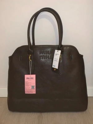 Brun handväska från Miu Miu i skinn - Säljer en brun handväska från Miu Miu i äkta skinn med präglad logga framtill och guldfärgade metalldetaljer. Väskan har dubbla handtag och en klassisk, stilren form. Perfekt för dig som gillar lyxiga accessoarer med exklusiv känsla.