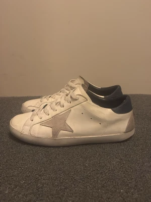 Golde Goose Skor - Säljer ett par vita sneakers i skinn med grå mockadetalj i form av en stjärna på sidan. Skorna har snörning och mörkblå hälkappa. Klassisk och clean look som passar till det mesta. Sulan är platt och tån är rund. (AI Beskrivning) Vid frågor så är det bara att skriva!