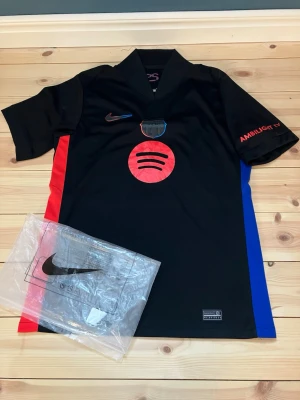 Svart Barcelona fotbollströja M - Säljer en svart Nike fotbollströja med röda och blå sidopaneler. Tröjan har Spotify-logga på bröstet, Ambilight-tryck på ärmen och UNHCR-logga bak. Tillverkad i Dri-FIT polyester för bästa komfort på planen. Perfekt för dig som älskar fotboll och vill ha en unik look.