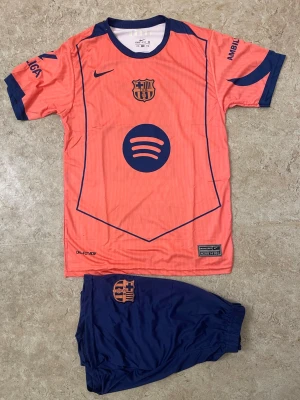 Barça Lamine Yamal matchställ Nike - Säljer ett FC Barcelona matchställ med Lamine Yamal 19-tryck. Tröjan är korallrosa med marinblå detaljer, klubbmärke och sponsorloggor. Shortsen är marinblå med klubbmärke. Tillverkad i lätt polyester, perfekt för fotbollsträning.