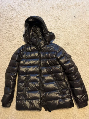 Svart Moncler pufferjacka med huva - Säljer en svart pufferjacka från Moncler med glansig finish och huva. Jackan har dragkedja framtill, två fickor med dragkedja och en extra ficka med dragkedja. Perfekt för kalla dagar och riktigt snygg till streetwear.