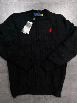 Svart stickad tröja från Polo Ralph Lauren - Svart kabelstickad tröja från Polo Ralph Lauren med klassisk rund hals och röd broderad logga på bröstet. Tröjan har långa ärmar och ribbade muddar vid ärmslut och nederkant. Perfekt för dig som gillar stilrena och tidlösa plagg.