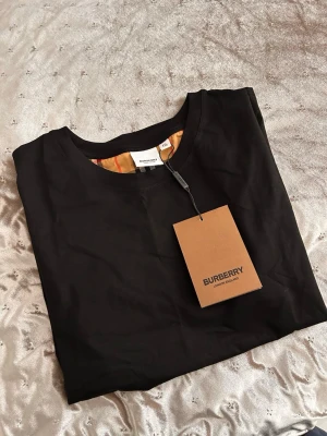 Burberry svart t-shirt med rutiga ärmar - Svart t-shirt från Burberry i storlek XXL men sitter som M. Klassiska rutiga ärmar i beige, rött och svart. T-shirten har rund halsringning och är tillverkad i mjuk bomull med lite elastan för extra komfort. Perfekt för dig som gillar stilrena plagg med en twist. STORLEK XXL MEN SITTER SOM M