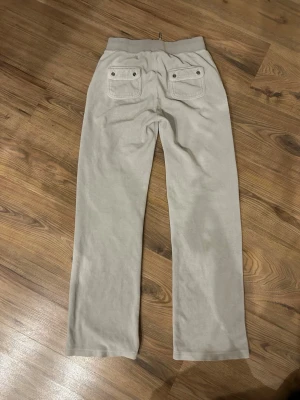 Grå mjukis set Juicy Couture - Säljer ett par beige mjukisbyxor från Juicy Couture med raka ben och bred resår i midjan. Byxorna har snörning och två bakfickor med knappdetaljer samt en broderad JC-logga på ena fickan. Tillverkade i mjuk velour som känns riktigt skön. + tröja