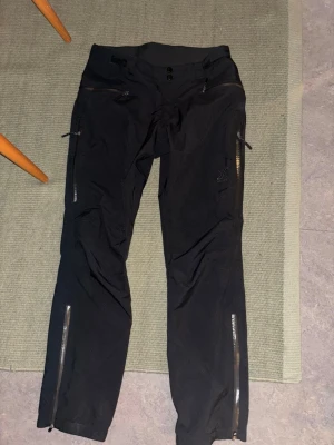 Svarta skalbyxor RevolutionRace dam M - Säljer ett par svarta skalbyxor från RevolutionRace i damstorlek M. Byxorna har flera dragkedjeförsedda fickor, vädring med nät vid låren, justerbar midja med kardborre och detaljerade sömmar. Perfekta för utomhusaktiviteter och har en clean look med RVRC-logga på benet. Modellen heter: Silence Proshell 3L Pants Dam Black/Anthracite Storlek: M