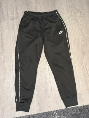 Svarta mjukisbyxor från Nike - Svarta mjukisbyxor från Nike med vit logga på vänster lår och vita detaljer längs sidorna. Byxorna har elastisk midja och muddar vid bensluten. Perfekta för chill eller träning, riktigt sköna och stilrena. Byxorna är i bra skick och dom har inga bakfickor.