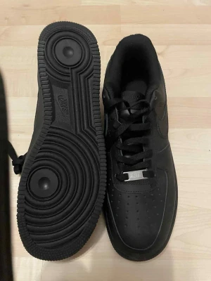 Svarta Nike Air Force 1 sneakers - Säljer ett par helsvarta Nike Air Force 1 sneakers i klassisk low modell. Skorna har en stilren design med perforerad tå, rund tå och snörning. Tillverkade i skinn med en rejäl platt sula och diskret Nike-logga på sidan. Perfekta för dig som gillar tidlös streetstil.