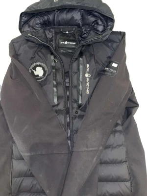 Svart dunjacka från Sail Racing - Säljer en svart dunjacka från Sail Racing med huva och snygga detaljer som patch på bröstet och logga på ärmen. Jackan har dragkedja framtill och quiltad design som ger en sportig vibe. Perfekt för kalla dagar när du vill hålla stilen. 