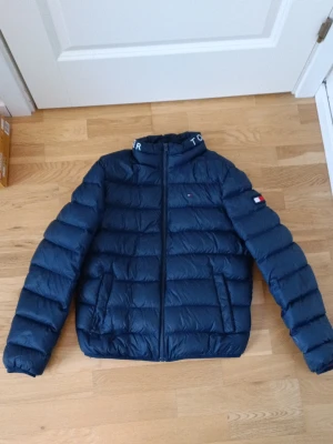 jacka Tommy Hilfiger storlek 164 - Säljer en mörkblå pufferjacka från Tommy Hilfiger med dragkedja och hög krage där loggan står broderad. Jackan har två fickor fram och röd insida. Klassisk Hilfiger-flagga på ärmen och liten logga på bröstet. Perfekt för kalla dagar. Jackan är i storlek 164.