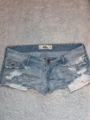 Ljusblå slitna jeansshorts Hollister - Säljer ett par ljusblå jeansshorts från Hollister med låg midja och slitna detaljer. Klassisk femficksmodell, råa kanter och broderad logga på fickan. Midjemått rakt över: 41 cm