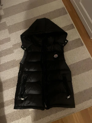 Svart dunväst från Moncler med huva - Säljer en svart, lång dunväst från Moncler med stor huva och två dragkedjeförsedda fickor framtill. Västen har quiltad design och klassisk Moncler-logga på bröstet. Perfekt för lager-på-lager och riktigt snygg till höst- och vinterlooken.