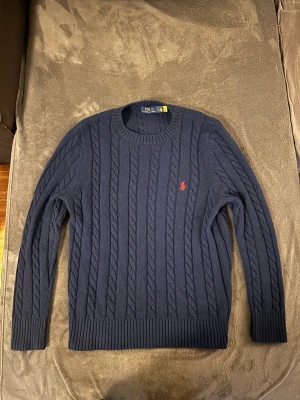 Mörkblå kabelstickad tröja Polo Ralph Lauren - Kabelstickad mörkblå tröja från Polo Ralph Lauren med klassisk röd broderad logga på bröstet. Tröjan har rund halsringning, ribbade muddar och är långärmad. Perfekt för dig som vill ha en tidlös och snygg look.