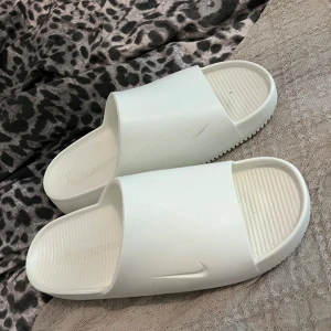 Vita Nike slides i syntetmaterial - Säljer ett par stilrena vita Nike slides i syntetmaterial. De har en enkel och clean design med öppen tå och diskret Nike-logga på sidan. Perfekta för sommaren eller att chilla hemma. Ribbad sula för extra grepp.