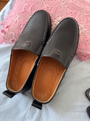 Svarta loafers i skinn, storlek 41 - Säljer ett par klassiska svarta loafers i skinn med dekorativa sömmar runt ovandelen. Skorna har en mjuk innersula och en rejäl, räfflad yttersula för bra grepp. Perfekta till både jeans och chinos för en clean look.