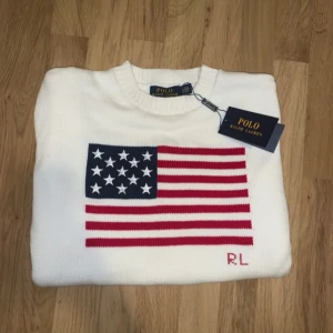 Vit stickad tröja Polo Ralph Lauren - Vit stickad tröja från Polo Ralph Lauren med amerikansk flagga i rött, blått och vitt på bröstet samt RL-broderi. Klassisk rund halsringning och ribbade muddar. Perfekt för dig som gillar preppy stil och vill sticka ut.