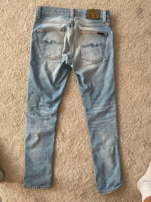 Ljusblå Nudie Jeans grim trim 28/32 - Säljer ett par ljusblå slim fit jeans från Nudie Jeans med klassisk femficksdesign och orangea sömmar. Jeansen har en snygg tvättad look och raka ben. Tillverkade i bomull för en skön känsla och stilren passform. Modellen är Grim trim. Jeansen är ljusblåa med en vit tvätt