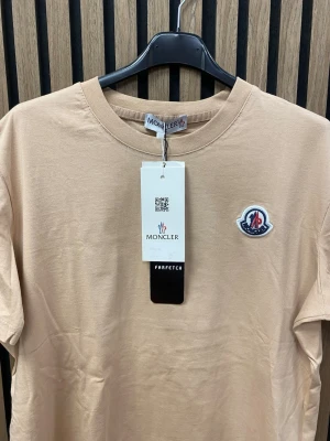 Beige t-shirt  - Snygg beige t-shirt klassisk rund hals och broderad logga på bröstet. Tillverkad i mjuk bomull för en skön känsla. Enkel och stilren design som passar perfekt till jeans eller shorts.