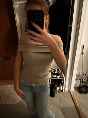 Beige offshoulder topp - Säljer en beige offshoulder topp med tight passform. Toppen är ärmlös och har en bred kant över axlarna som ger en snygg och stilren look. Perfekt att matcha med jeans eller kjol för en trendig outfit.