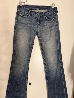 Blå bootcut jeans från 7 For All Mankind - Snygga blå jeans från 7 For All Mankind med bootcut-snitt och klassisk femficksdesign. Jeansen har en tvättad look med ljusare partier och detaljerade sömmar. De är Lågmidjade och vintage, köpta secondhand! Storlek 26