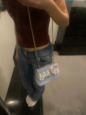 Blå axelväska med blommor och kedja - Säljer en ljusblå axelväska i tweedliknande material med guldfärgad kedjerem. Väskan har dekorativa blommor i vitt och en stor tygblomma i beige och grått på framsidan samt ett vridlås i guld. Finns små defekter! Pris kan diskuteras 💗