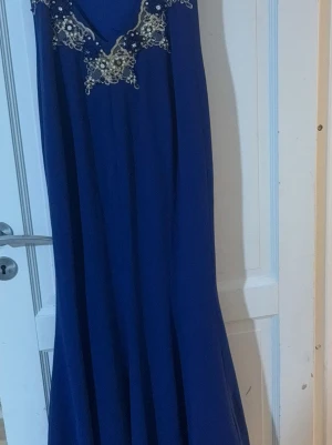 Blå långklänning med pärldetaljer - Elegant blå långklänning med v-ringning och vackra pärl- och spetsdetaljer vid bysten. Klänningen har en flytande passform och är ärmlös, perfekt för bal eller fest. Materialet känns lätt och faller snyggt längs kroppen.
