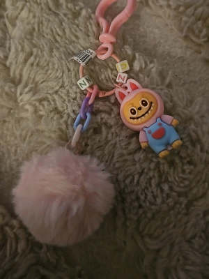 Rosa fluffig nyckelring med figur - Söt nyckelring med en stor, fluffig rosa boll och en färgglad figur med kaninöron och blå overall. Kedjan har pastellfärgade länkar och bokstavspärlor. Perfekt att hänga på väskan eller nycklarna för en lekfull touch.