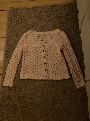 Ljusrosa stickad kofta från ellos - Supersöt ljusrosa stickad kofta från Ellos med genombrutet mönster och bruna knappar framtill. Koftan har lång ärm, ribbade muddar och en croppad passform som ger en trendig look. Perfekt att slänga över en topp för extra stilpoäng.