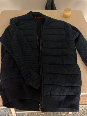 Svart quiltad kofta från Armor Lux - Säljer en svart quiltad kofta från Armor Lux i storlek M. Koftan har ribbade ärmar och sidor, samt dragkedja framtill. Materialet känns mjukt och värmande, perfekt för kyligare dagar. Enkel och stilren design som funkar till det mesta.