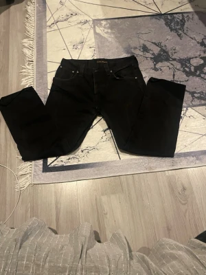 Svarta jeans från Nudie Jeans - Svarta jeans från Nudie Jeans med klassisk femficksdesign och silverfärgade nitar. Jeansen har rak passform och är tillverkade i slitstark bomull. Snyggt broderad logga på insidan och knapp med märkets namn. Perfekt för dig som gillar stilrena svarta jeans.