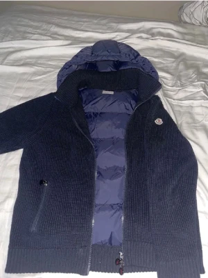 Mörkblå stickad Moncler pufferjacka - Säljer en mörkblå stickad pufferjacka från Moncler med quiltad insida och huva. Jackan har dragkedja framtill, ribbade muddar och en Moncler-logga på bröstet. Perfekt för kyliga dagar och riktigt snygg till jeans eller sweatpants.