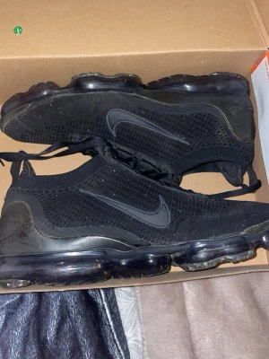 Nike Vapormax Flyknit Svarta US11 - Säljer ett par svarta Nike Vapormax Flyknit med stickad ovandel och genomskinlig sula. Skorna har snörning och den klassiska Nike-loggan på sidan. Superlätta och flexibla med en modern look och bekväm passform.