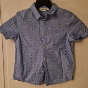 Blå prickig skjorta från H&M - Kortärmad skjorta från H&M i ljusblått med små mörkblå prickar. Klassisk krage och knäppning framtill med vita knappar. Tillverkad i mjuk bomull som känns skön mot huden. Hund finns i hemmet. Kolla in mina andra annonser för samfrakt! 