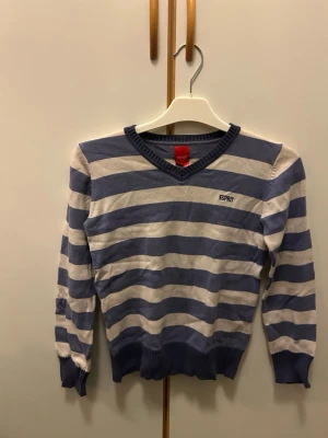 Randig lila och vit tröja från Esprit - Snygg stickad tröja från Esprit med breda lila och vita ränder. V-ringad hals och ribbade muddar vid ärmslut och nederkant. Perfekt för dig som gillar klassisk stil med en twist. Esprit-logga broderad på bröstet. Passar dig som vill ha en bekväm och stilren look.