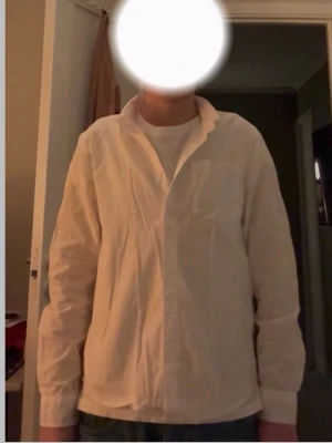 Vit skjorta med klassisk krage - Säljer en stilren vit skjorta med klassisk krage och lång ärm. Skjortan har en bröstficka och knappar framtill. Passformen är normal och materialet känns som bomull, vilket gör den skön att bära. Perfekt för dig som gillar enkel och clean stil passar dig som är 157 cm 