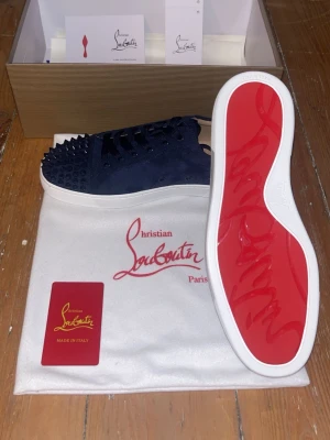 Christian loubutin - Säljer ett par mörkblå sneakers från Christian Louboutin med coola nitar på tån och sidorna. Skorna har snörning, vit sula och röd undersida. Tillverkade i mocka med detaljer i skinn på insidan. Perfekt för dig som gillar att sticka ut med din stil.