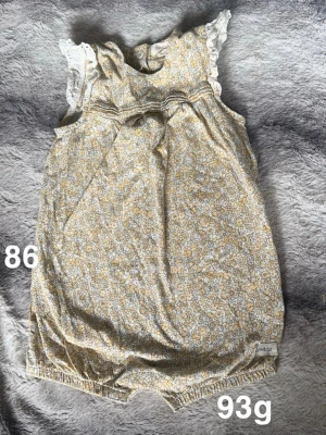 Blommig byxdress från Newbie - Supersöt byxdress från Newbie i ljusgul och vit med blommigt mönster. Volangdetaljer vid axlarna och knappar i nacken. resår vid benen för skön passform. Perfekt för varma dagar!🌸 Fint använt skick. Storlek 86 