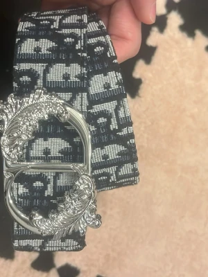 Svart och vit mönstrad bälte med silverdetaljer - Snyggt bälte med svart och vit mönster och en stor, dekorerad silverspänne. Bältet har ett grafiskt mönster och är tillverkat i syntetmaterial med inslag av textil. Perfekt för att ge outfiten en extra edge och cool vibe.