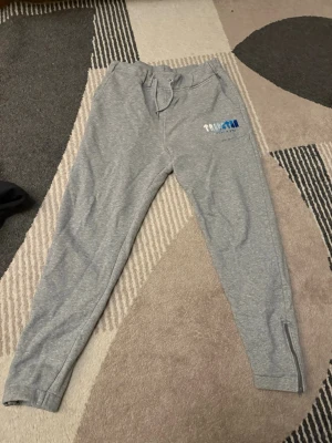 Grå mjukisbyxor från Trapstar - Säljer ett par grå mjukisbyxor från Trapstar med broderad logga i olika blå nyanser på vänster lår. Byxorna har snörning i midjan och dragkedja vid bensluten. Perfekta för chill eller träning, riktigt sköna och stilrena.