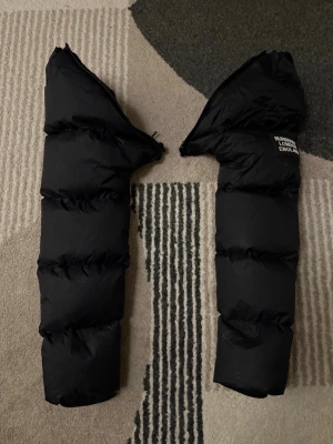 Svarta pufferjacka ärmar Burberry - Säljer två svarta, tjocka och quiltade ärmar från Burberry pufferjacka med märkets logga på ena ärmen. Perfekt för dig som vill byta ut eller laga din jacka. Materialet är syntet och dun, och designen är stilren och modern.