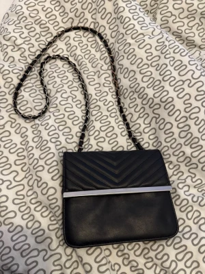 Svart quiltad axelväska med kedja - Snygg svart axelväska med quiltad ovandel och silverfärgad metallist framtill. Väskan har en lång kedjerem med insydda skinnband för en cool look. Perfekt storlek för det viktigaste och passar till många outfits.
