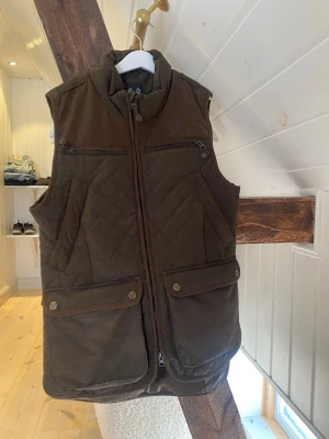 Brun quiltad väst från Vapiti S - Säljer en brun quiltad väst från jägarliv i storlek S. Västen har flera fickor med dragkedja och tryckknappar, hög krage och snyggt rutmönster. Perfekt för lager-på-lager och jakt. Materialet är slitstarkt och känns robust.