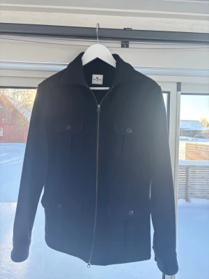 Mörkblå overshirt från John Henric - Säljer en mörkblå overshirt från John Henric med fyra fickor framtill och dragkedja. Jackan har klassisk krage och är tillverkad i ull, vilket gör den både stilren och varm. Perfekt lager-på-lager-plagg för kallare vårdagar.