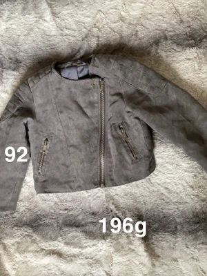 Grå mockaimitations jacka barn - Snygg grå jacka i mockaimitation med coola detaljer som sned dragkedja och två fickor med dragkedjor. Jackan har quiltade axelpartier och är perfekt för dig som vill ha en tuff look. 🌸 Fint använt skick. Storlek 92