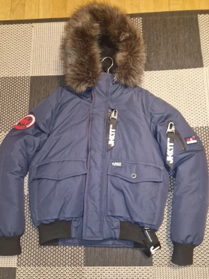 Blå vinterjacka från JAK1T med pälshuva - Säljer en mörkblå pufferjacka från JAK1T med stor, fluffig pälskantad huva och feta loggdetaljer. Jackan har flera fickor med lock, ribbade muddar och dragkedja framtill. Perfekt för kalla dagar och riktigt snygg streetstil. obs..äkta dunjacka !!!